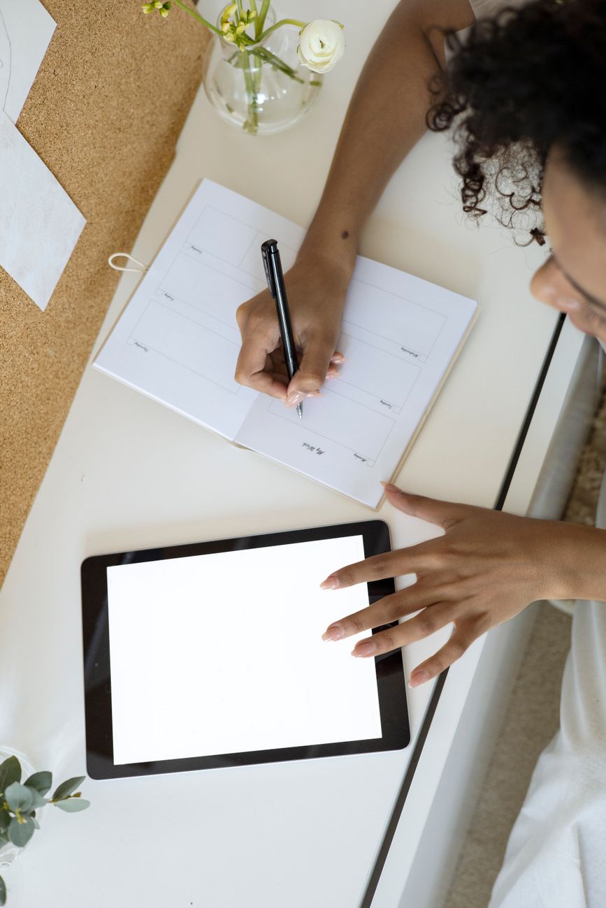 Hands writing in a paper planner next to a tablet